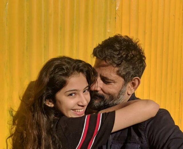 Sara Arjun smiling and hugging a bearded man in front of a yellow background.